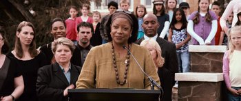 Movie still from “Knowing” (2009), directed by Alex Proyas – A woman is speaking at a podium in front of a group of people; Medium shot, Over the shoulder angle
