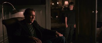 Movie still from “Knowing” (2009), directed by Alex Proyas – A man sitting on a couch next to a boy; Medium shot, Over the shoulder angle