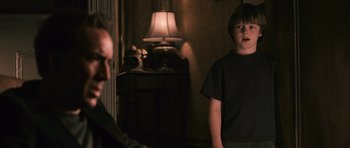 Movie still from “Knowing” (2009), directed by Alex Proyas – A young boy standing next to a man in front of a lamp; Medium shot, Over the shoulder angle
