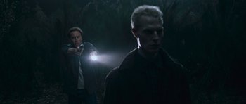 Movie still from “Knowing” (2009), directed by Alex Proyas – A man holding a flashlight while standing next to another man in a dark forest; Medium shot, Over the shoulder angle