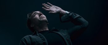 Movie still from “Knowing” (2009), directed by Alex Proyas – A man in a black shirt and black shirt holding his hand up to his face; Close Up shot, Low angle