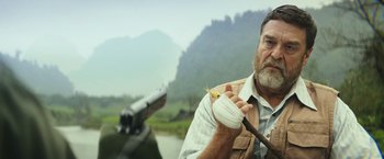 Movie still from “Kong: Skull Island” (2017), directed by Jordan Vogt-Roberts – A man with a bird in his hand and a gun in the background; Close Up shot, Low angle