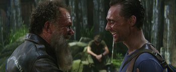 Movie still from “Kong: Skull Island” (2017), directed by Jordan Vogt-Roberts – Two men are laughing together in the woods; Medium shot, Over the shoulder angle