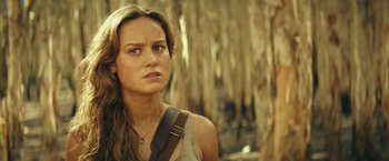 Movie still from “Kong: Skull Island” (2017), directed by Jordan Vogt-Roberts – A person wearing a tank top near a forest; Close Up shot, Over the shoulder angle