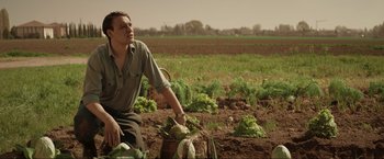 Movie still from “Lamborghini: The Man Behind the Legend” (2022), directed by Bobby Moresco – A man sitting in a field holding a basket of vegetables; Medium shot, Low angle