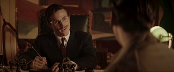 Movie still from “Lamborghini: The Man Behind the Legend” (2022), directed by Bobby Moresco – A man sitting at a table talking to a woman in a suit; Medium shot, Over the shoulder angle