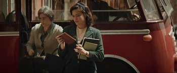Movie still from “Lamborghini: The Man Behind the Legend” (2022), directed by Bobby Moresco – A woman is reading a book while standing in front of a bus; Medium shot, Over the shoulder angle