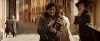 Movie still from “Lamborghini: The Man Behind the Legend” (2022), directed by Bobby Moresco – A woman in a brown coat is holding a book; Medium shot, Over the shoulder angle