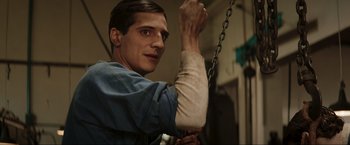 Movie still from “Lamborghini: The Man Behind the Legend” (2022), directed by Bobby Moresco – A man holding onto a metal chain while wearing a blue shirt; Close Up shot, Low angle