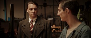 Movie still from “Lamborghini: The Man Behind the Legend” (2022), directed by Bobby Moresco – A man wearing a suit and tie standing next to another man; Medium shot, Over the shoulder angle