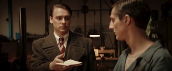 Movie still from “Lamborghini: The Man Behind the Legend” (2022), directed by Bobby Moresco – A man wearing a suit and tie holding a book; Medium shot, Over the shoulder angle