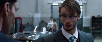 Movie still from “Lamborghini: The Man Behind the Legend” (2022), directed by Bobby Moresco – A man in a suit and tie standing in front of some cars; Close Up shot, Over the shoulder angle