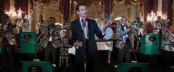 Movie still from “Lamborghini: The Man Behind the Legend” (2022), directed by Bobby Moresco – A man in a suit and tie holding a microphone; Medium shot, Low angle