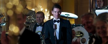 Movie still from “Lamborghini: The Man Behind the Legend” (2022), directed by Bobby Moresco – A man in a tuxedo standing in front of a microphone; Medium shot, Over the shoulder angle