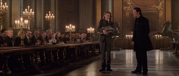 Movie still from “Lara Croft: Tomb Raider” (2001), directed by Simon West – A man standing in front of a long table holding books; Wide shot, Low angle
