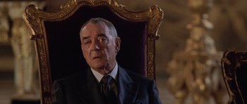 Movie still from “Lara Croft: Tomb Raider” (2001), directed by Simon West – An older man wearing a suit and tie sitting in a chair; Close Up shot, Over the shoulder angle