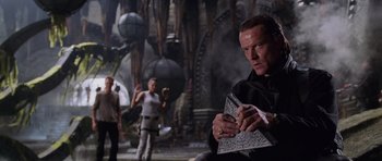 Movie still from “Lara Croft: Tomb Raider” (2001), directed by Simon West – A man sitting on the ground holding a book; Medium shot, Over the shoulder angle