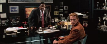 Movie still from “Last Action Hero” (1993), directed by John McTiernan – Two men are sitting at a desk in a room; Medium shot, High angle