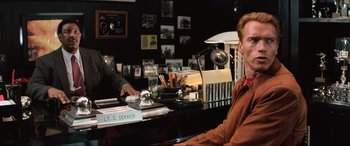Movie still from “Last Action Hero” (1993), directed by John McTiernan – A man sitting at a desk in front of a laptop; Medium shot, Over the shoulder angle