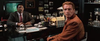 Movie still from “Last Action Hero” (1993), directed by John McTiernan – A man sitting at a desk in front of a wall of pictures; Medium shot, Over the shoulder angle