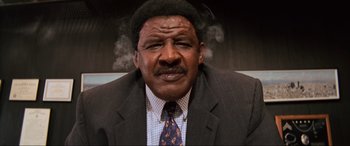 Movie still from “Last Action Hero” (1993), directed by John McTiernan – An older man in a suit and tie smoking a cigarette; Close Up shot, Low angle