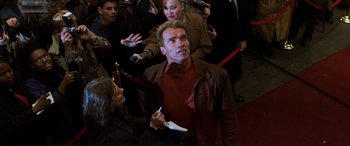 Movie still from “Last Action Hero” (1993), directed by John McTiernan – A man in a red shirt and a brown jacket is surrounded by reporters; Medium shot, High angle