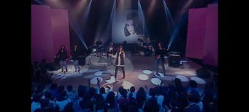 Movie still from “Laura Pausini: Pleasure to Meet You” (2022), directed by Ivan Cotroneo – A group of people on a stage singing; Extreme Wide shot, High angle