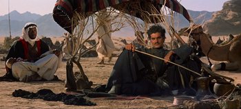 Movie still from “Lawrence of Arabia” (1962), directed by David Lean – A man sitting on the ground holding a stick; Wide shot, Low angle