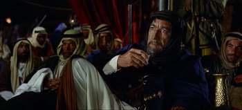 Movie still from “Lawrence of Arabia” (1962), directed by David Lean – An old man holding a cigarette in front of a group of men; Medium shot, Over the shoulder angle