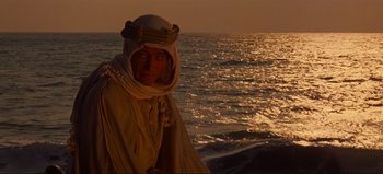 Movie still from “Lawrence of Arabia” (1962), directed by David Lean – A man wearing a turban standing in front of a body of water at sunset; Medium shot, Over the shoulder angle