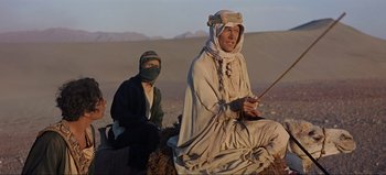 Movie still from “Lawrence of Arabia” (1962), directed by David Lean – A man sitting on top of a rock next to another man holding a stick; Medium shot, Low angle