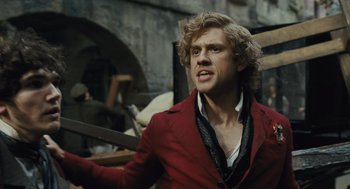 Movie still from “Les Misérables” (2012), directed by Tom Hooper – A man in a red jacket is standing in front of a brick wall; Medium shot, Low angle