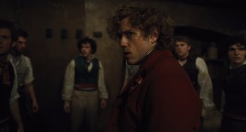 Movie still from “Les Misérables” (2012), directed by Tom Hooper – A man in a red jacket standing next to two other men; Medium shot, Over the shoulder angle