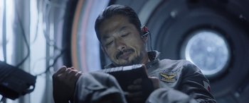 Movie still from “Life” (2017), directed by Daniel Espinosa – A man with headphones on looking at a book; Close Up shot, Over the shoulder angle
