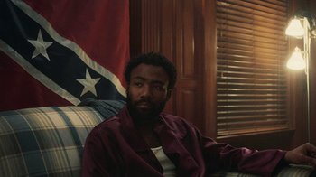 Movie still from “Atlanta” (2016), created by Donald Glover – A man sitting on top of a couch in front of a flag; Medium shot, Over the shoulder angle
