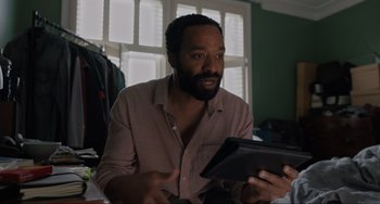 Movie still from “Locked Down” (2021), directed by Doug Liman – A man is sitting at a table and holding a tablet computer in his hands; Close Up shot, Low angle