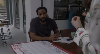 Movie still from “Locked Down” (2021), directed by Doug Liman – A man sitting at a wooden table in front of papers; Medium shot, Over the shoulder angle
