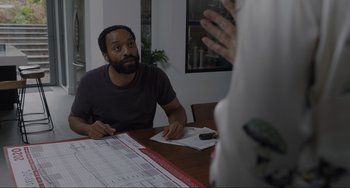 Movie still from “Locked Down” (2021), directed by Doug Liman – A man sitting at a table with papers on the table; Medium shot, Over the shoulder angle