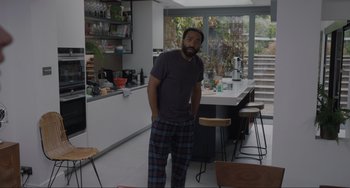 Movie still from “Locked Down” (2021), directed by Doug Liman – A man standing in a kitchen with his hands in his pockets; Medium shot, Over the shoulder angle