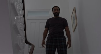 Movie still from “Locked Down” (2021), directed by Doug Liman – A man standing in front of a staircase wearing pajamas; Medium shot, Low angle