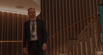 Movie still from “Locked Down” (2021), directed by Doug Liman – A man in a suit and tie standing in front of a staircase; Medium shot, Over the shoulder angle