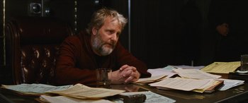 Movie still from “Looper” (2012), directed by Rian Johnson – A man sitting at a table with papers and a glass of water; Close Up shot, Over the shoulder angle