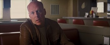 Movie still from “Looper” (2012), directed by Rian Johnson – A man sitting at a table in front of a window; Close Up shot, Over the shoulder angle
