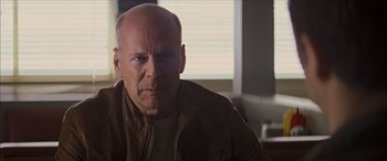 Movie still from “Looper” (2012), directed by Rian Johnson – A bald man with a goatee is sitting in front of a window; Close Up shot, Over the shoulder angle