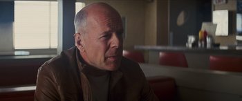 Movie still from “Looper” (2012), directed by Rian Johnson – A man with a bald head sitting in front of a window; Close Up shot, Over the shoulder angle