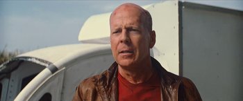 Movie still from “Looper” (2012), directed by Rian Johnson – A bald man in a red shirt and a brown jacket; Close Up shot, Low angle