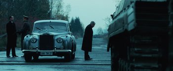 Movie still from “Lord of War” (2005), directed by Andrew Niccol – An older man standing in the middle of a road next to an old car; Wide shot, Low angle