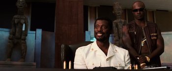 Movie still from “Lord of War” (2005), directed by Andrew Niccol – A man sitting at a table in front of a wooden statue; Close Up shot, Low angle