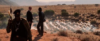 Movie still from “Lord of War” (2005), directed by Andrew Niccol – Two men standing on the side of a road near tents; Wide shot, Over the shoulder angle