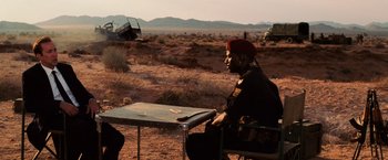 Movie still from “Lord of War” (2005), directed by Andrew Niccol – A man sitting at a table in the middle of the desert; Medium shot, Over the shoulder angle